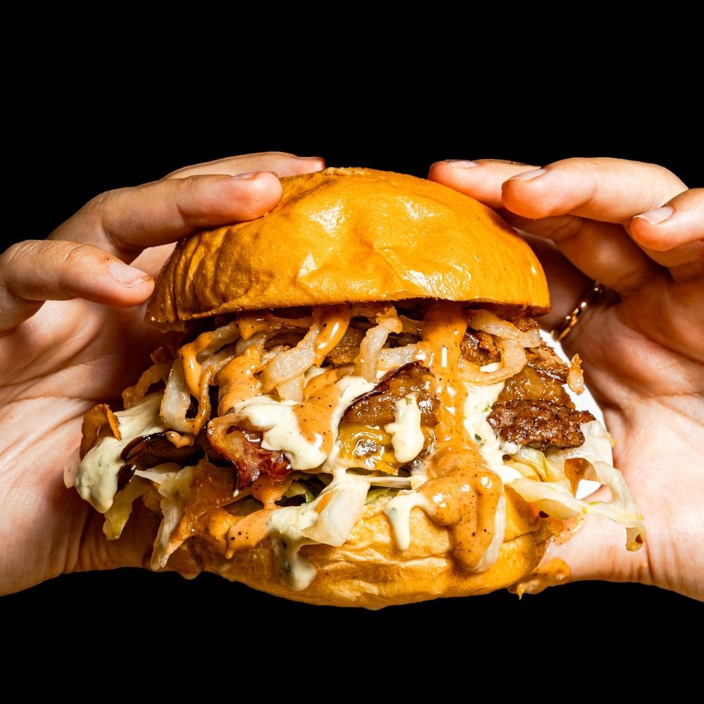 Una hamburguesa doble sostenida con las dos manos sobre fondo negro.