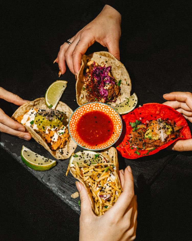 Cuatro manos cogen tacos a la vez alrededor de un cuenco de salsa roja.