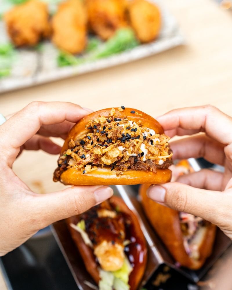 Dos manos sostienen un bao frito relleno de carne desmechada y cebolla crujiente.