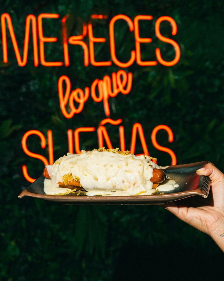 Un hot dog cubierto de salsa de queso, delante del neón naranja de la sala.