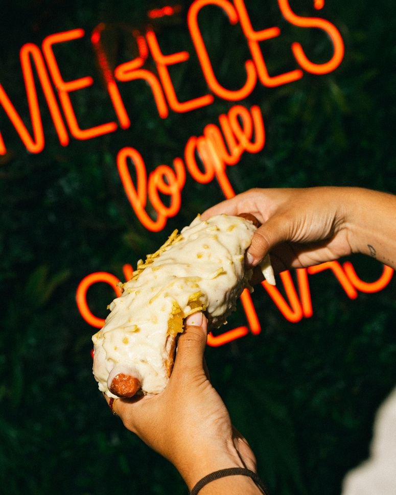 Una mano sostiene un burrito frito ante la pared de plantas y el neón «Mereces lo que sueñas».